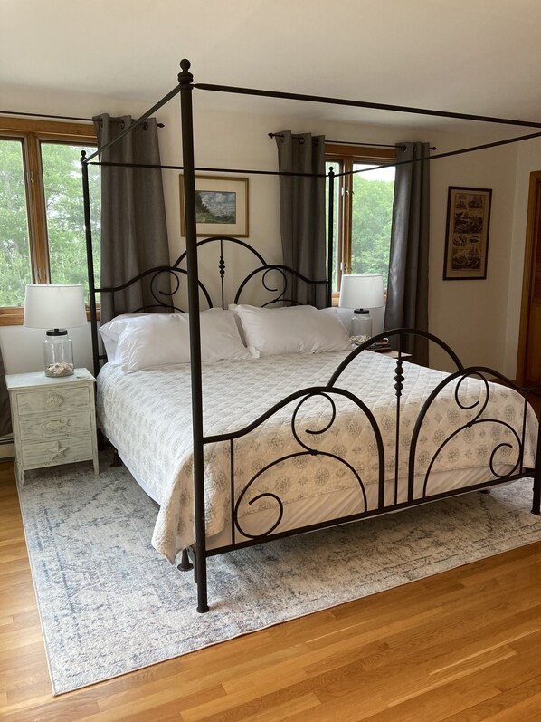 A four-poster bed with a white bedspread, two bedside tables with lamps, a rug, and a view of greenery through large windows.