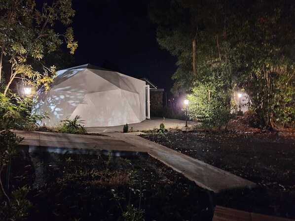 Un edificio moderno e geometrico con una superficie riflettente, circondato da alberi e illuminato da lampioni.