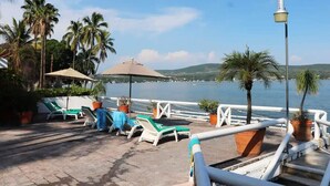 A deck with lounge chairs and umbrellas overlooking a body of water, surrounded by palm trees. - Incredibly Beautiful Big Villa For Up To 32 Guests (Tequesquitengo)