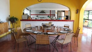 A dining area with a round table set for six, surrounded by wicker chairs. A kitchen is visible through an archway in the background. - Incredibly Beautiful Big Villa For Up To 32 Guests (Tequesquitengo)
