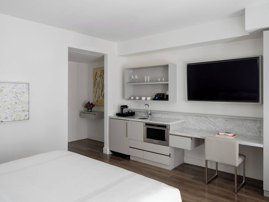 A modern hotel room with a bed, a desk, a TV, a kitchen area with a sink and cabinets, and a painting on the wall.