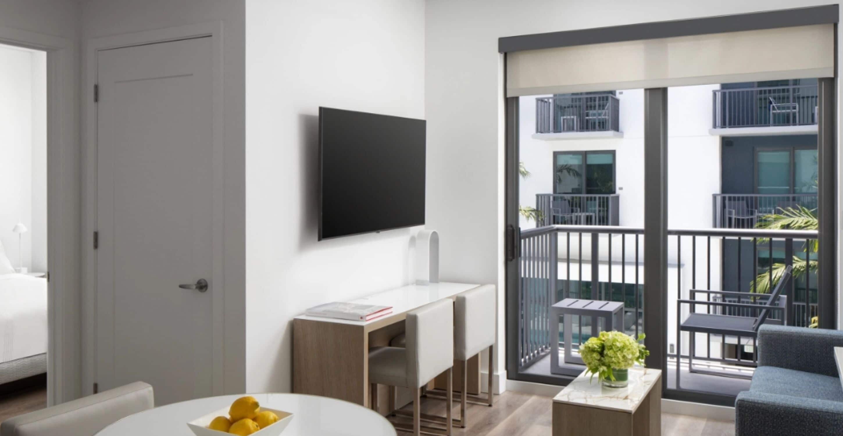A modern apartment with a flat-screen TV mounted on the wall, a dining area with a table and chairs, and a balcony with a view of the building's exterior.