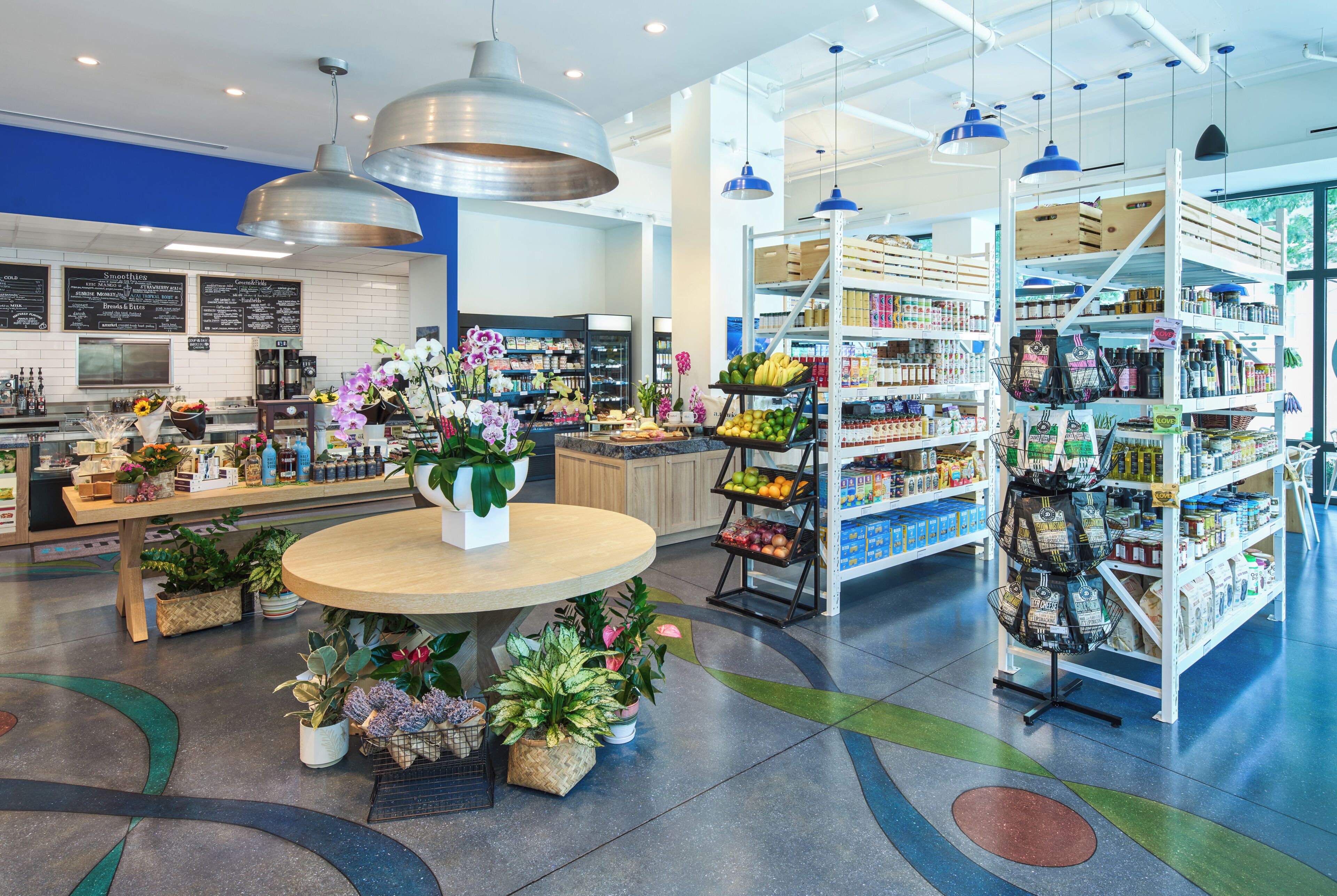 A modern grocery store with a central round table, potted plants, and shelves stocked with various products.