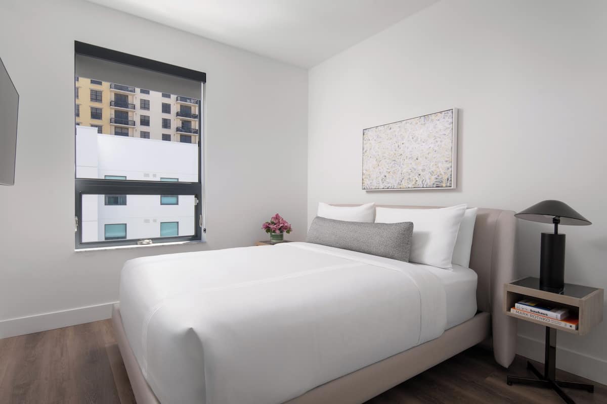 Een moderne slaapkamer met een groot bed, een raam met uitzicht op gebouwen, een nachtkastje met een lamp en een kunstwerk in een lijst aan de muur.