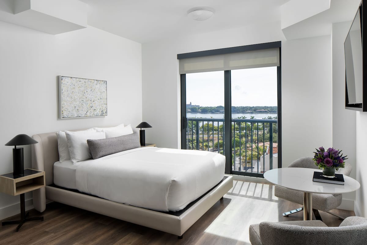 Een moderne slaapkamer met een bed, nachtkastjes, een klein rond tafeltje met een vaas, en een balkon met uitzicht.
