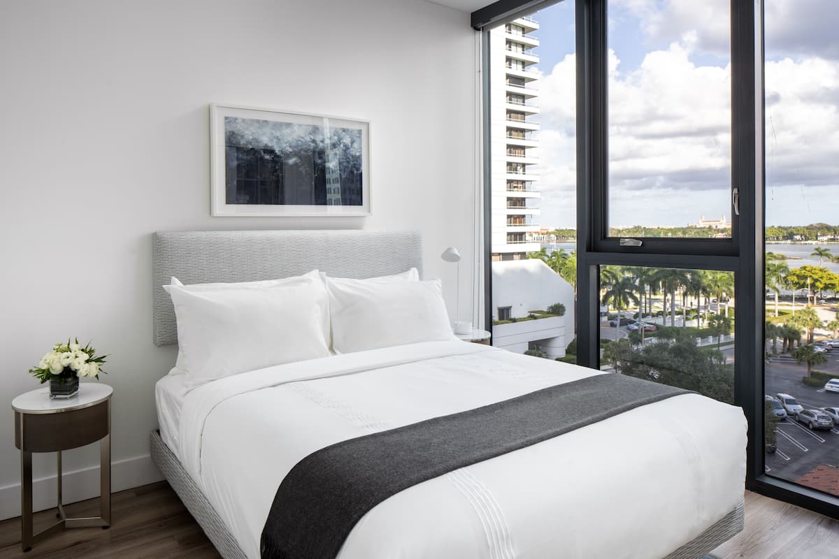 Een moderne hotelkamer met een groot bed, een bijzettafeltje met een bloevas, een ingelijste foto aan de muur en een stadsgezicht door een groot raam.