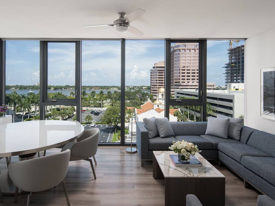 A modern living room with a large window, a ceiling fan, a dining area with chairs, and a sofa with cushions.
