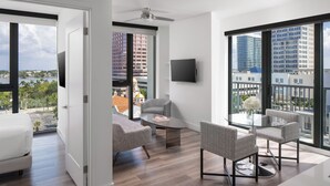 A modern hotel room with a large window, a flat-screen TV mounted on the wall, a comfortable seating area with a glass coffee table, and a bed with a white headboard.