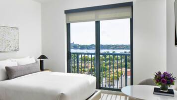 Habitación de hotel con una cama grande, una mesita con un florero y un balcón con vistas a la ciudad.