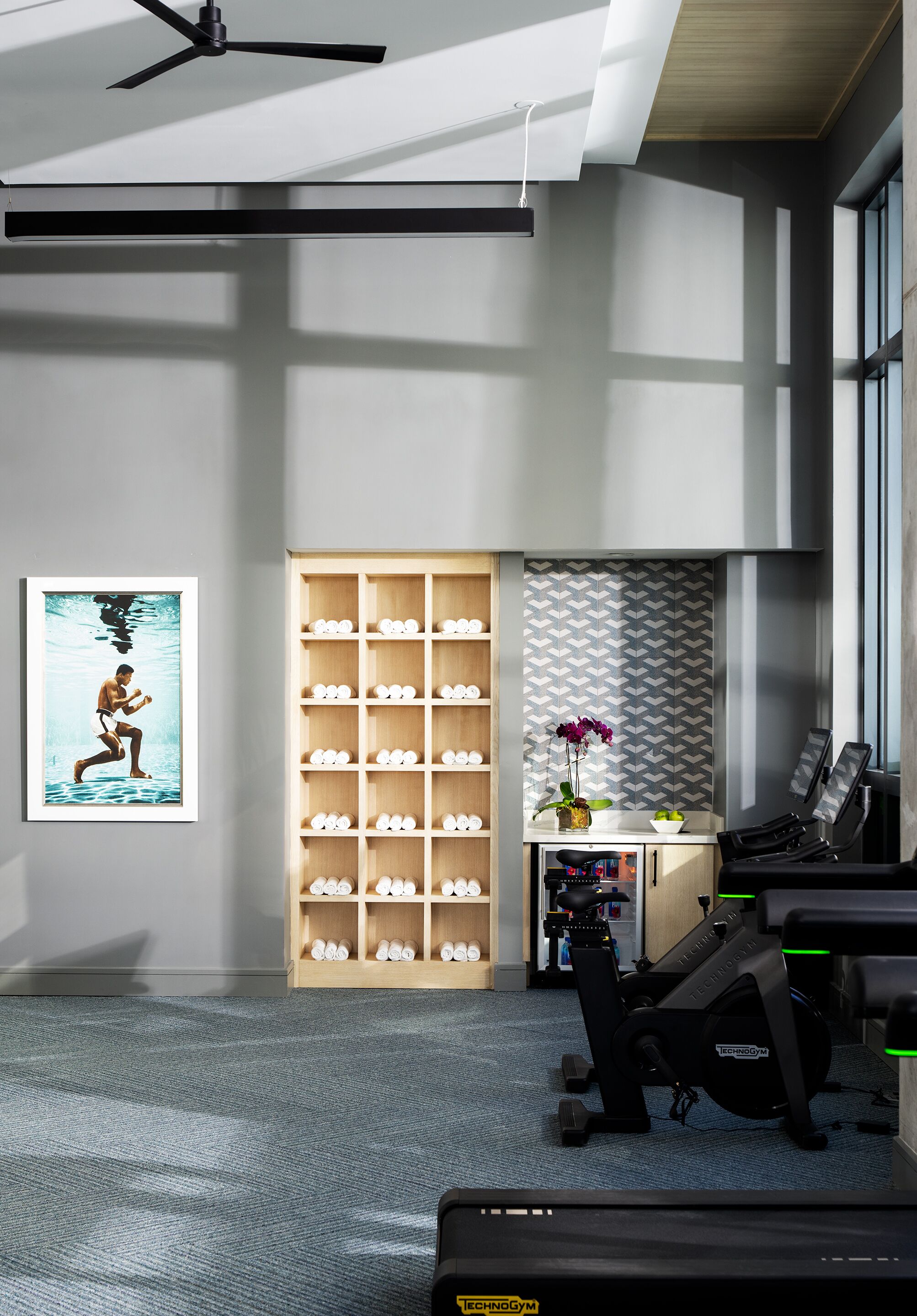 Un gimnasio moderno con equipo de ejercicio, un ventilador fijado a la pared y un estante con mancuernas ordenadas.