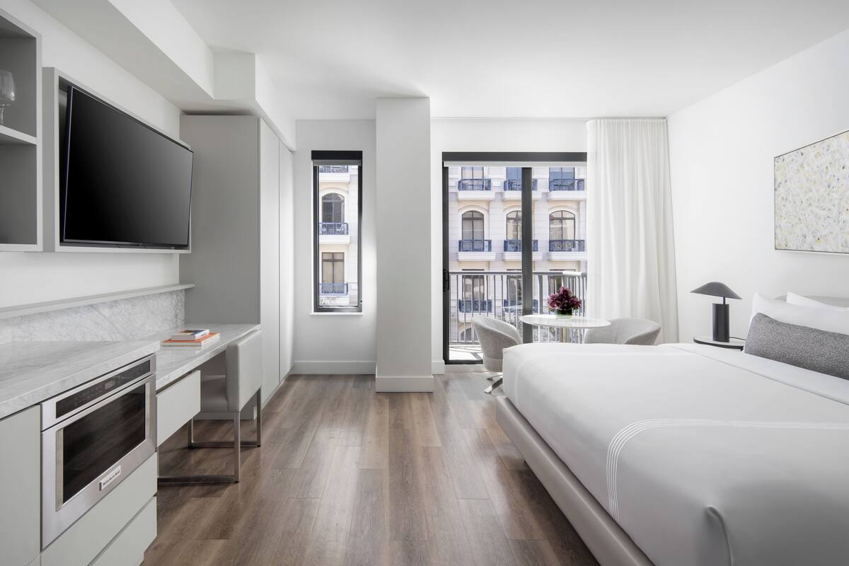 Een moderne hotelkamer met een groot bed, een bureau met een flatscreen-tv, een kleine tafel met een lamp en uitzicht op de stad door grote ramen.