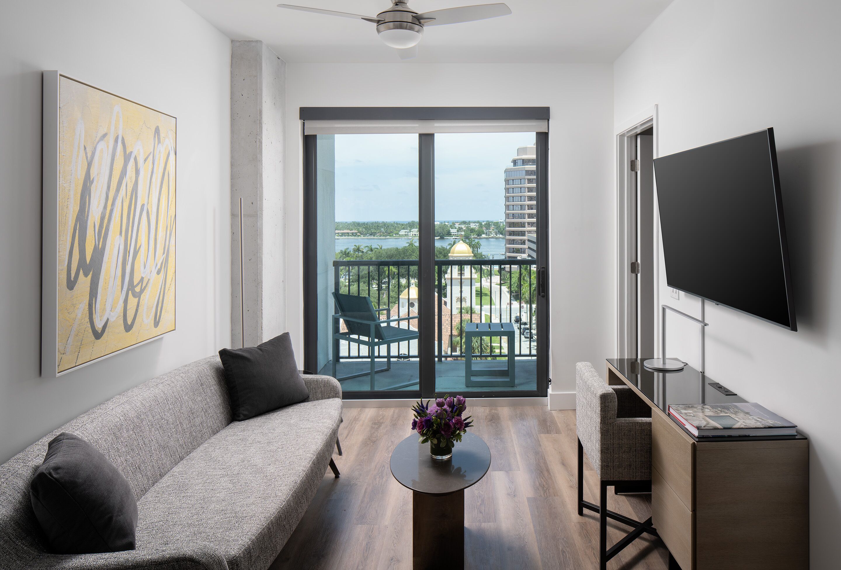 En moderne stue med en grå sofa, et sofabord med blomster, et fjernsyn på et stativ og en balkon med udsigt over byen og vandet.