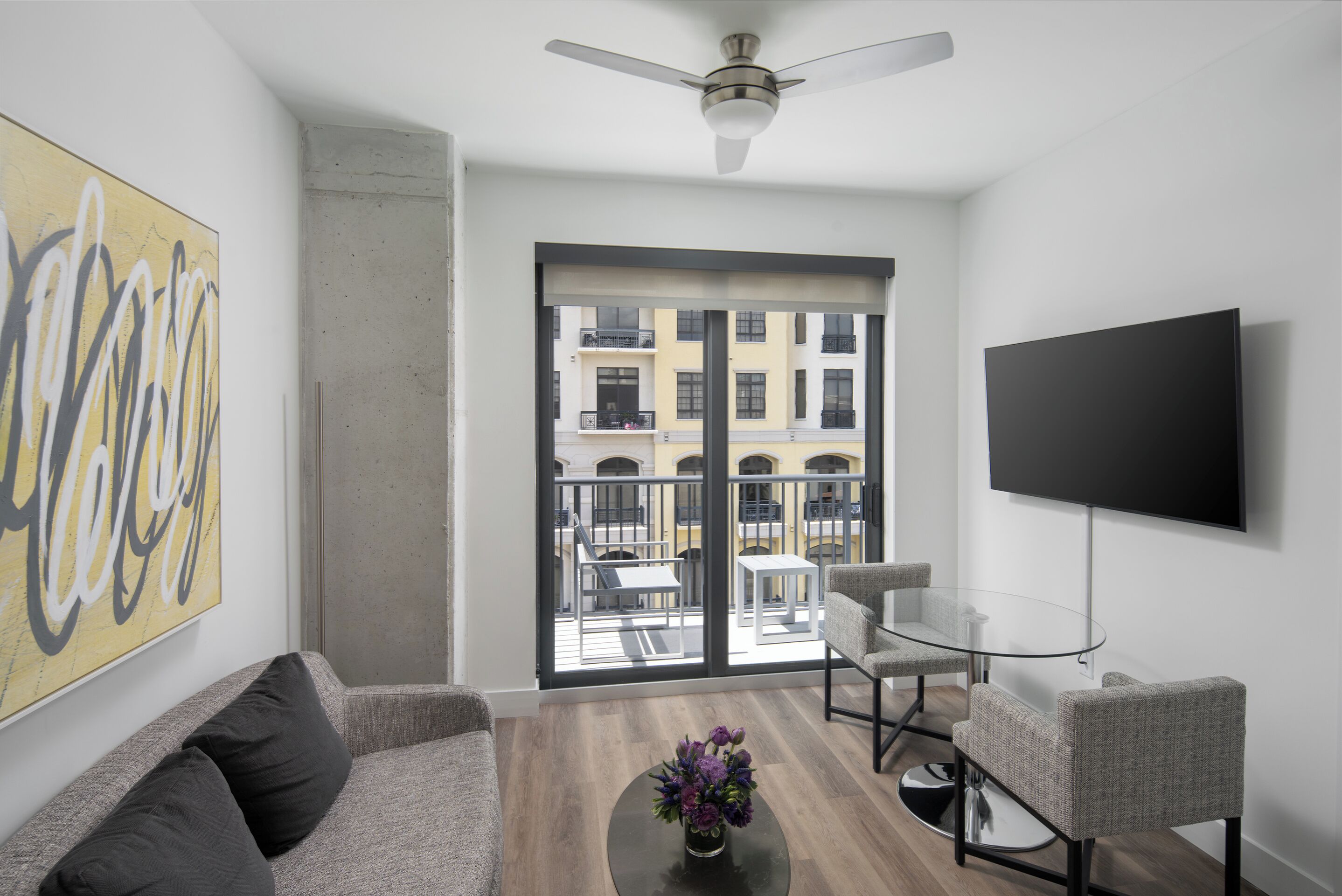 Un salon moderne comprenant un canapé, une table basse avec un vase, une table en verre avec des chaises, un téléviseur fixé au mur et un balcon avec du mobilier d’extérieur.