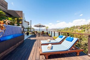 Une terrasse en bois avec deux chaises longues à rayures et une table, entourée de plantes en pot et offrant une vue sur un paysage vallonné.