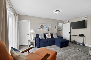 A living room with a sectional sofa, a TV mounted on the wall, and a small side table with a lamp.
