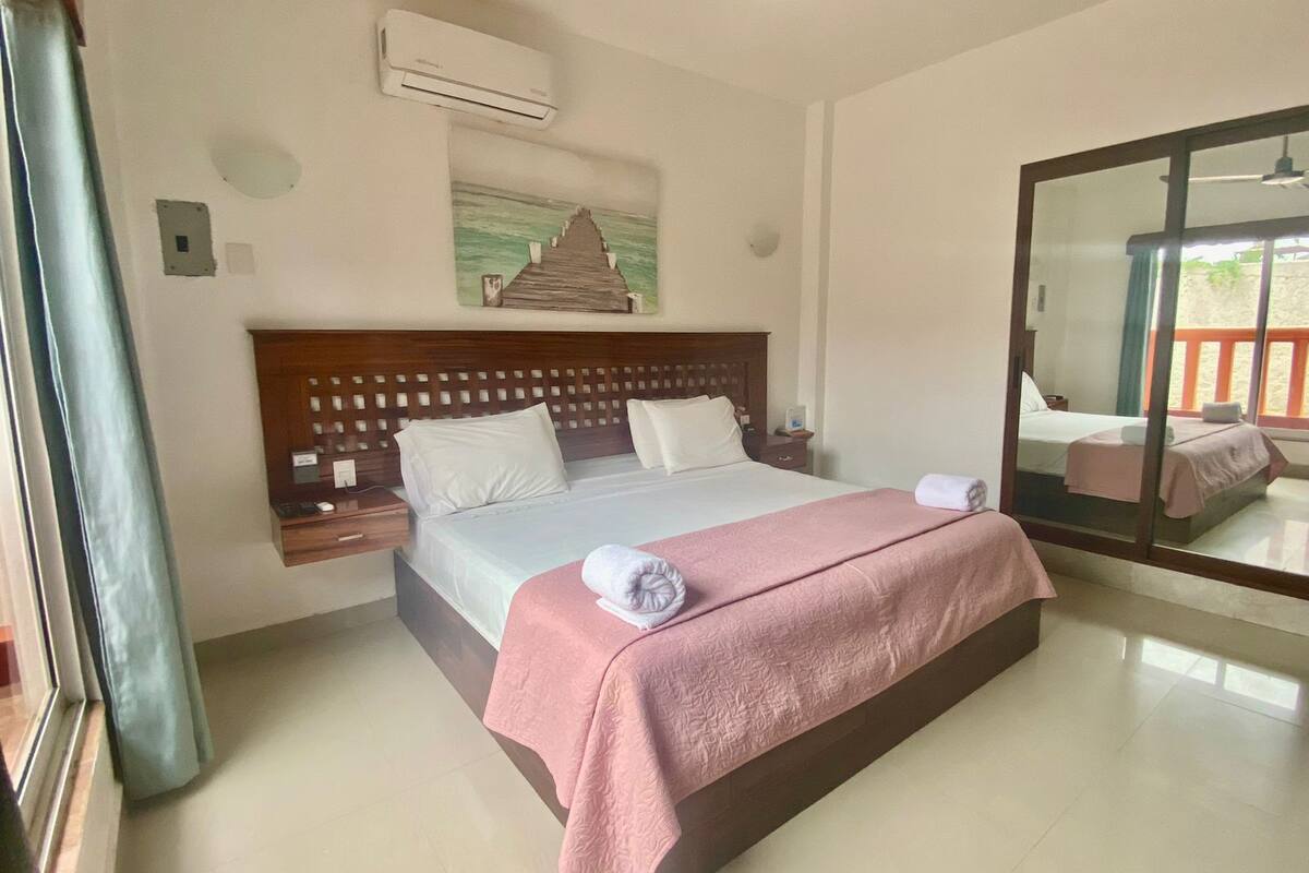 Un dormitorio amplio y bien iluminado, con una cama grande, cabecera de madera, mesita de noche y velador. Se observa un espejo grande en una pared, y un equipo de aire acondicionado instalado sobre la cama.