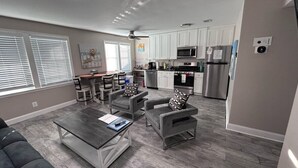 A modern living room with a grey sofa, two armchairs, and a coffee table. There is a kitchen in the background with white cabinets, a refrigerator, and a microwave.