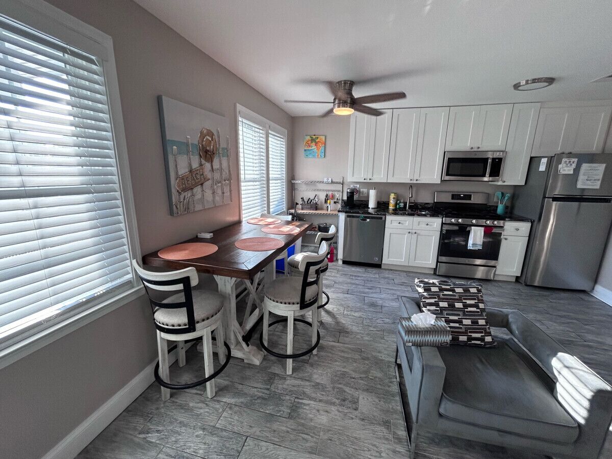 Una cocina moderna con armarios blancos, un comedor con mesa de madera y cuatro sillas, y un sofá gris en la sala.