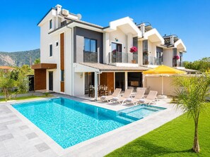 A modern two-story house with a swimming pool, outdoor seating, and a mountain backdrop.