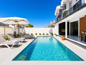 A rectangular swimming pool with lounge chairs and umbrellas, surrounded by a modern building with balconies and a clear blue sky.