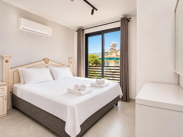 A hotel room with a bed, a nightstand, an air conditioning unit, a window with curtains, and a view of buildings outside.