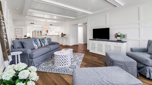 Un salon spacieux avec un canapé gris, des poufs et un téléviseur posé sur un meuble blanc.
