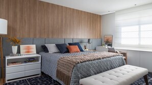 A modern bedroom with a wooden headboard, a bed with multiple pillows, a bedside table with a lamp, and a window with blinds. - Casa 1 (Rio de Janeiro)