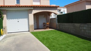 Una casa de dos pisos con garaje, un pequeño patio con una silla y un césped bien cuidado.