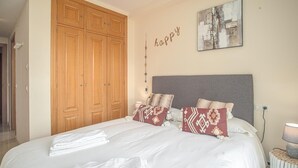 Een net opgemaakt bed met witte lakens en decoratieve kussens, een houten garderobe en een aan de muur hangend kunstwerk met de tekst 'happy'.