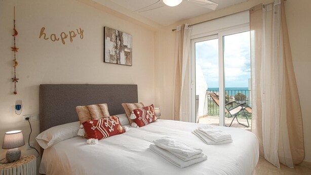 Een netjes opgemaakt bed met witte lakens, een grijs hoofdbord en decoratieve kussens. Daarboven hangt een kunstwerk aan de muur en staat een bordje met de tekst 'happy'.