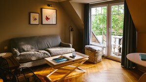 Un salon chaleureux avec un canapé gris, une table basse en bois, un fauteuil noir et une petite table avec une lampe. Des tableaux encadrés ornent le mur, et une grande fenêtre offre une vue sur l’extérieur.