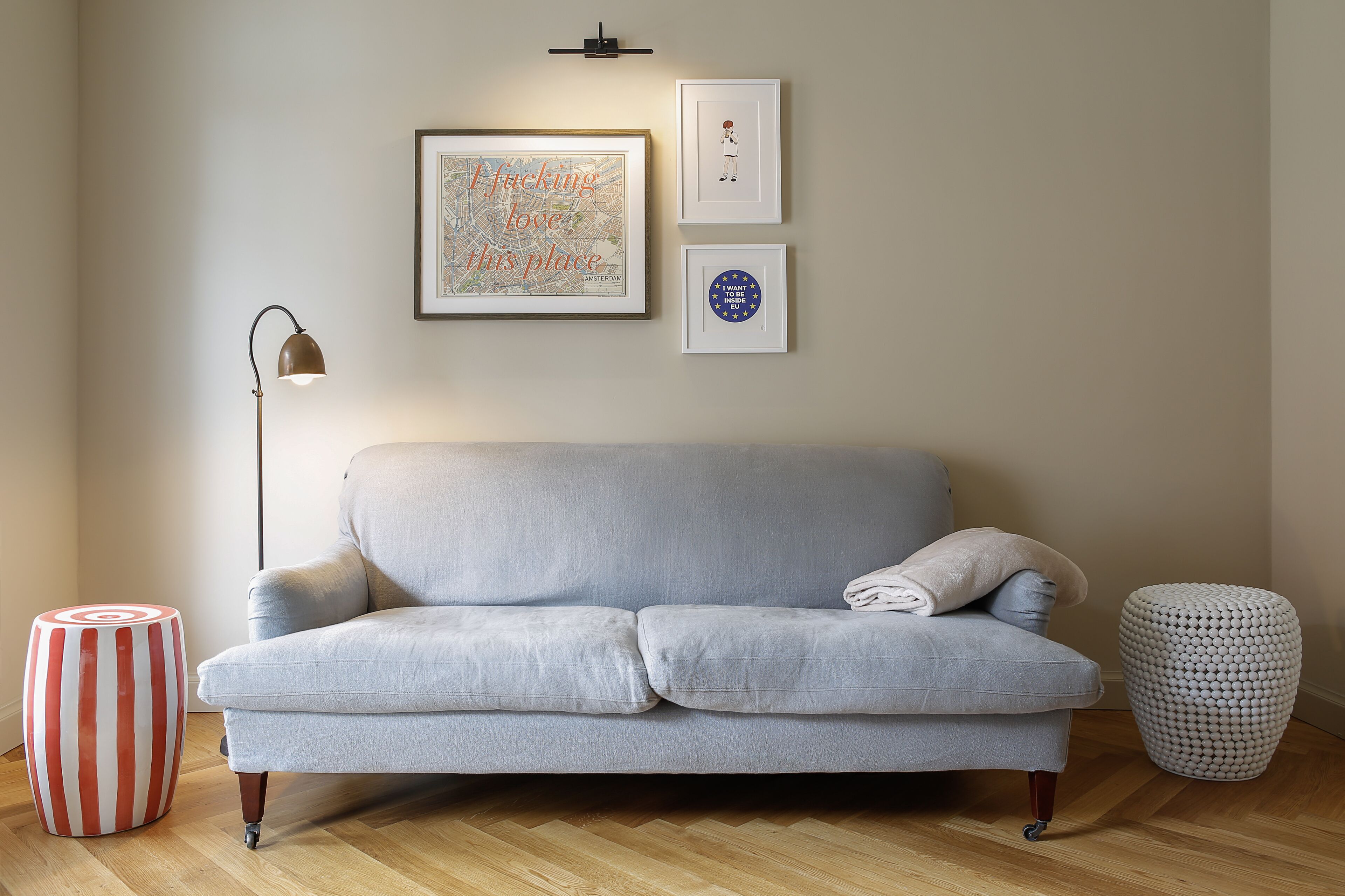 Un sofá de gris claro con un solo cojín, un reposapiés blanco y rojo a rayas, un taburete redondo blanco, y un mapa enmarcado con texto en la pared.