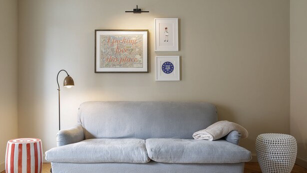 Un sofá de gris claro con un solo cojín, un reposapiés blanco y rojo a rayas, un taburete redondo blanco, y un mapa enmarcado con texto en la pared.