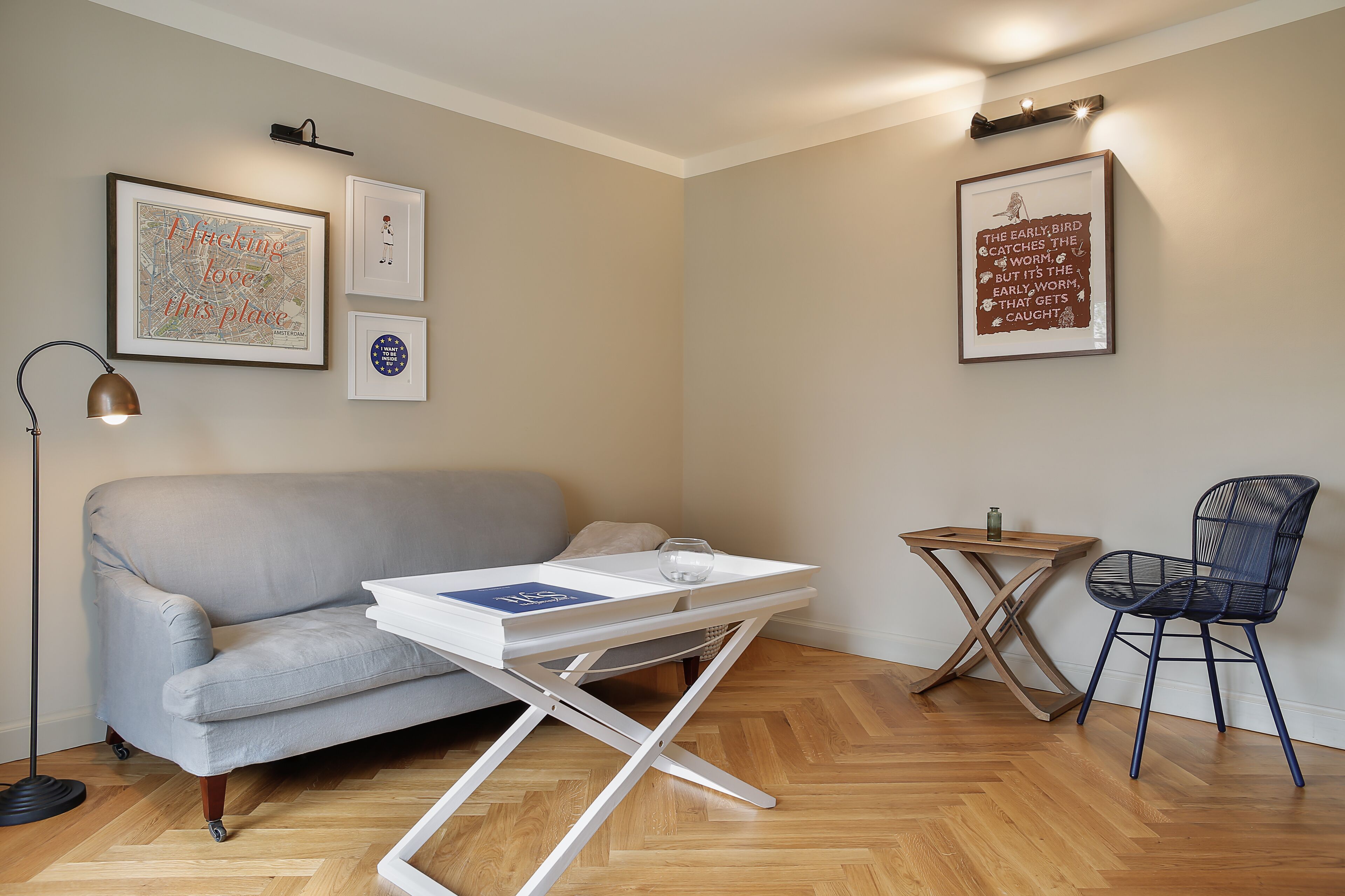 A modern living room with a grey sofa, a white coffee table, a blue chair, and a small wooden side table. There are framed artworks on the walls and a floor lamp.