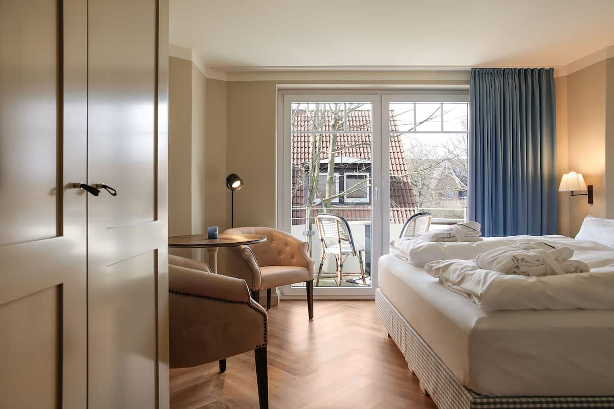 Quarto de hotel com cama, poltrona, mesinha, luminária e vista para o exterior.