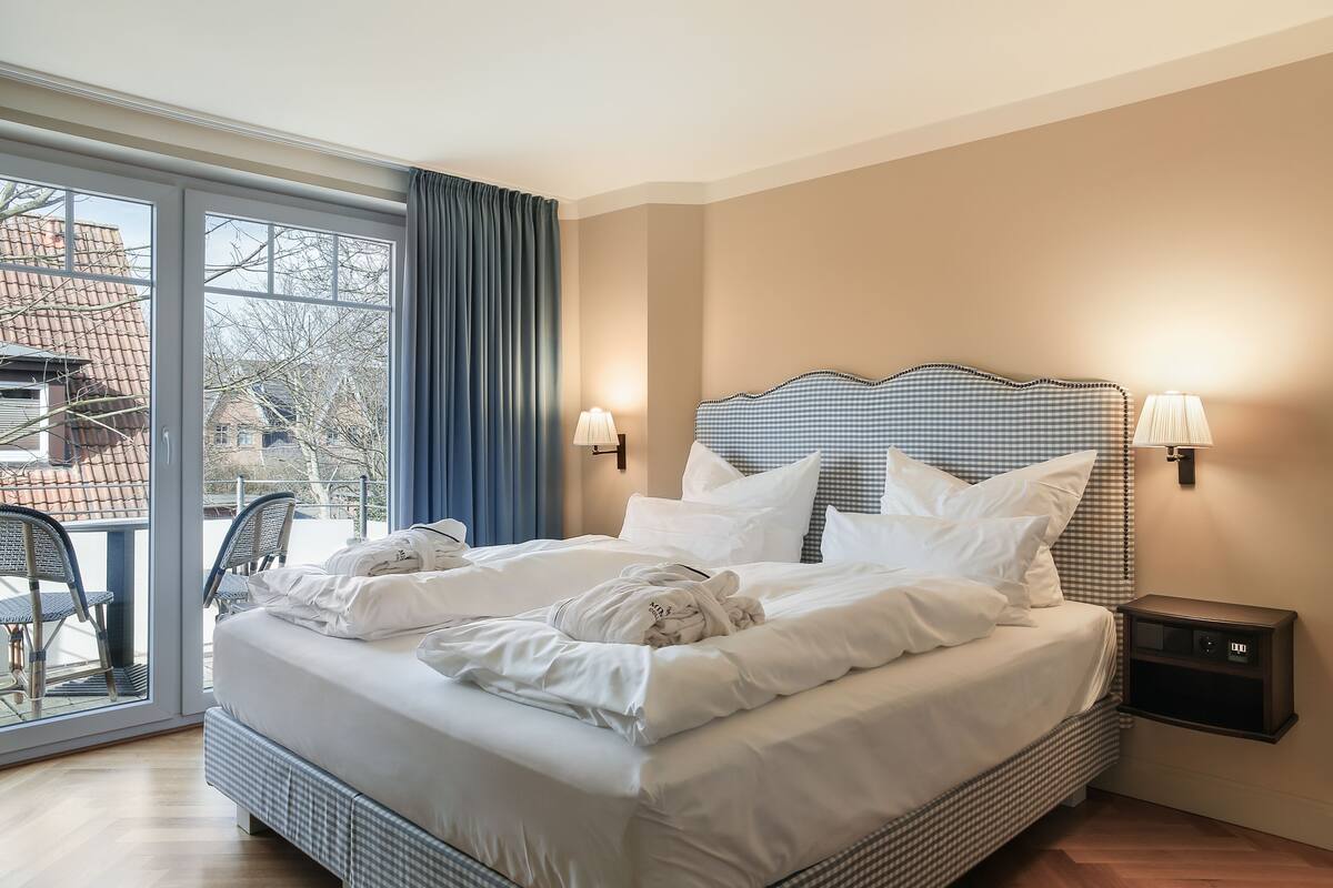 Quarto de hotel com uma cama grande, duas luminárias de cabeceira, uma mesa de cabeceira e vista através de uma porta de vidro.