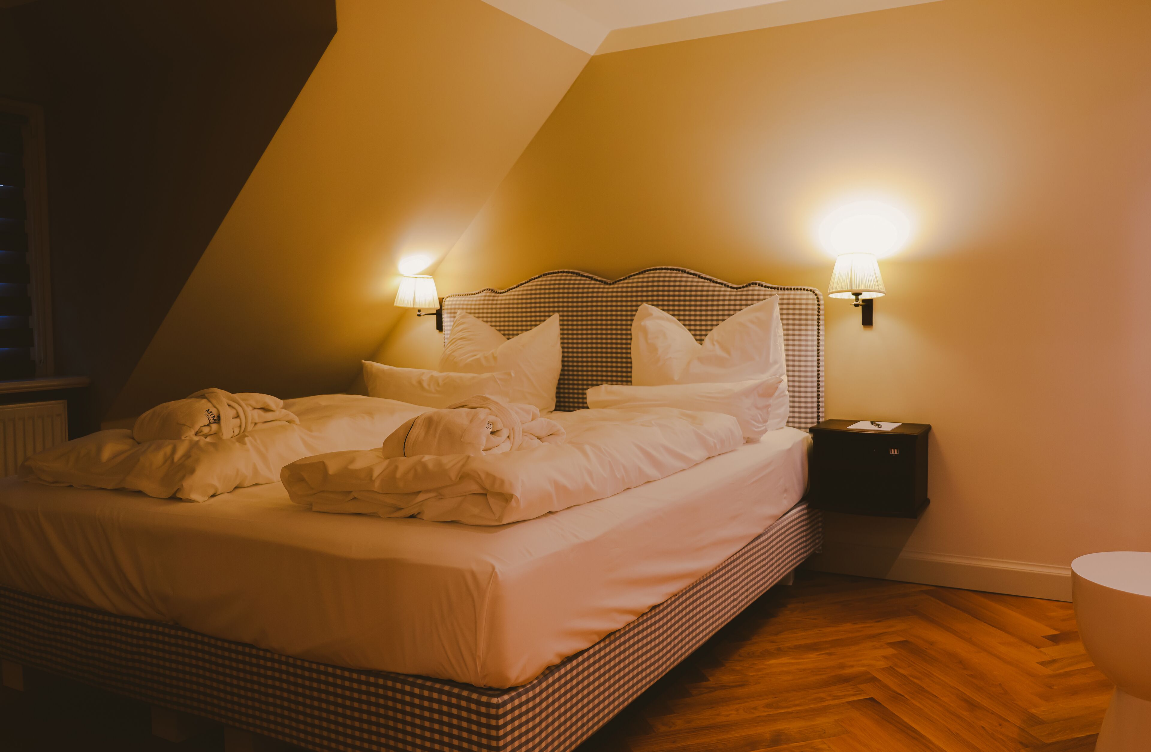 A neatly made bed with white linens and pillows, two wall-mounted lamps, a small bedside table, and a wooden floor.