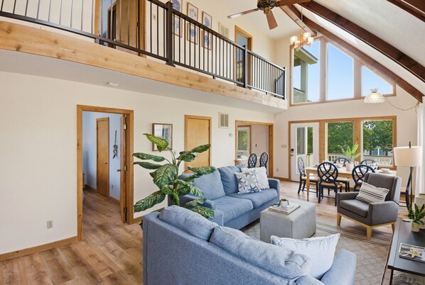 A spacious living area with a high ceiling, wooden beams, a balcony, and a dining area with a table and chairs.