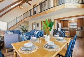 A dining area with a wooden table set for four, blue chairs, a staircase, and a kitchen in the background.