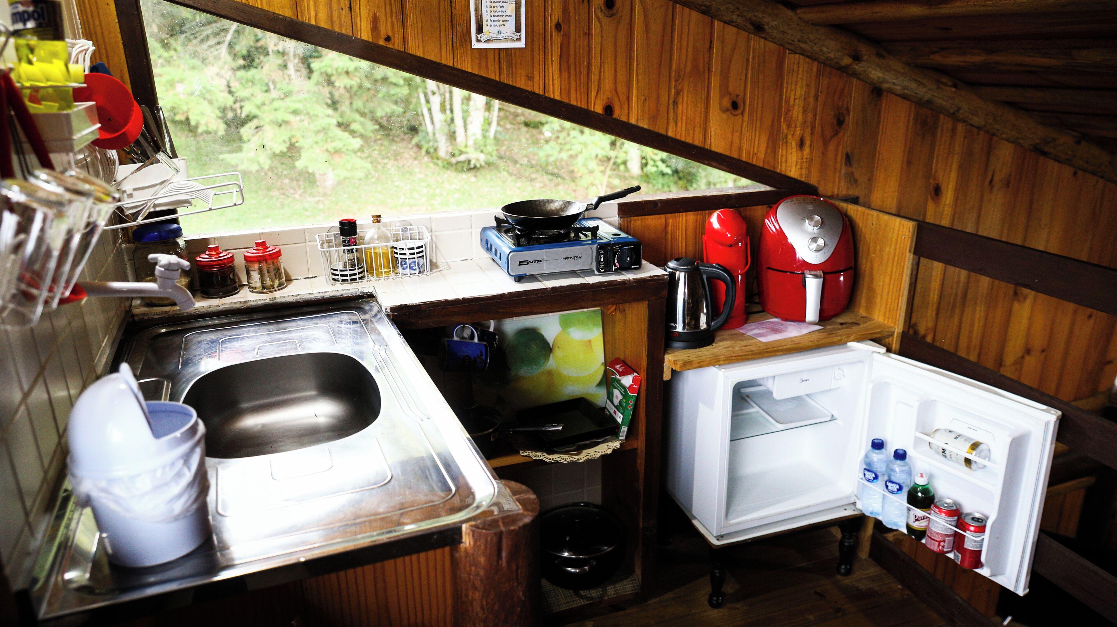 A compact kitchen with a sink, stove, and refrigerator, wooden walls, and a window overlooking greenery.