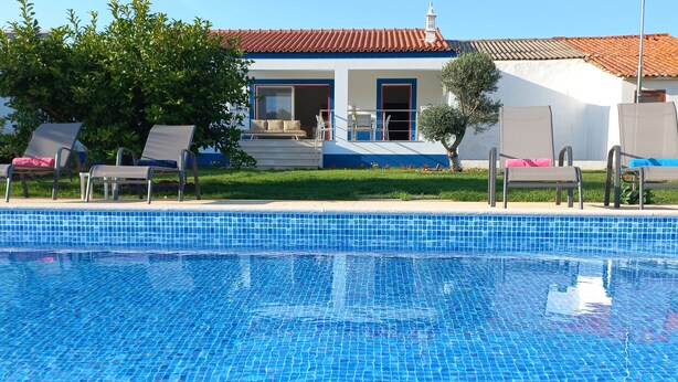 Une piscine aux carreaux bleu clair, un espace extérieur couvert avec des fauteuils de jardin, et un bâtiment avec un toit recouvert de tuiles rouges en arrière-plan.