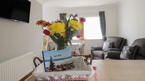 Een woonkamer met een aan de muur bevestigde televisie, een tafel met een 'Guest Book' en een mand, en een raam met gordijnen.