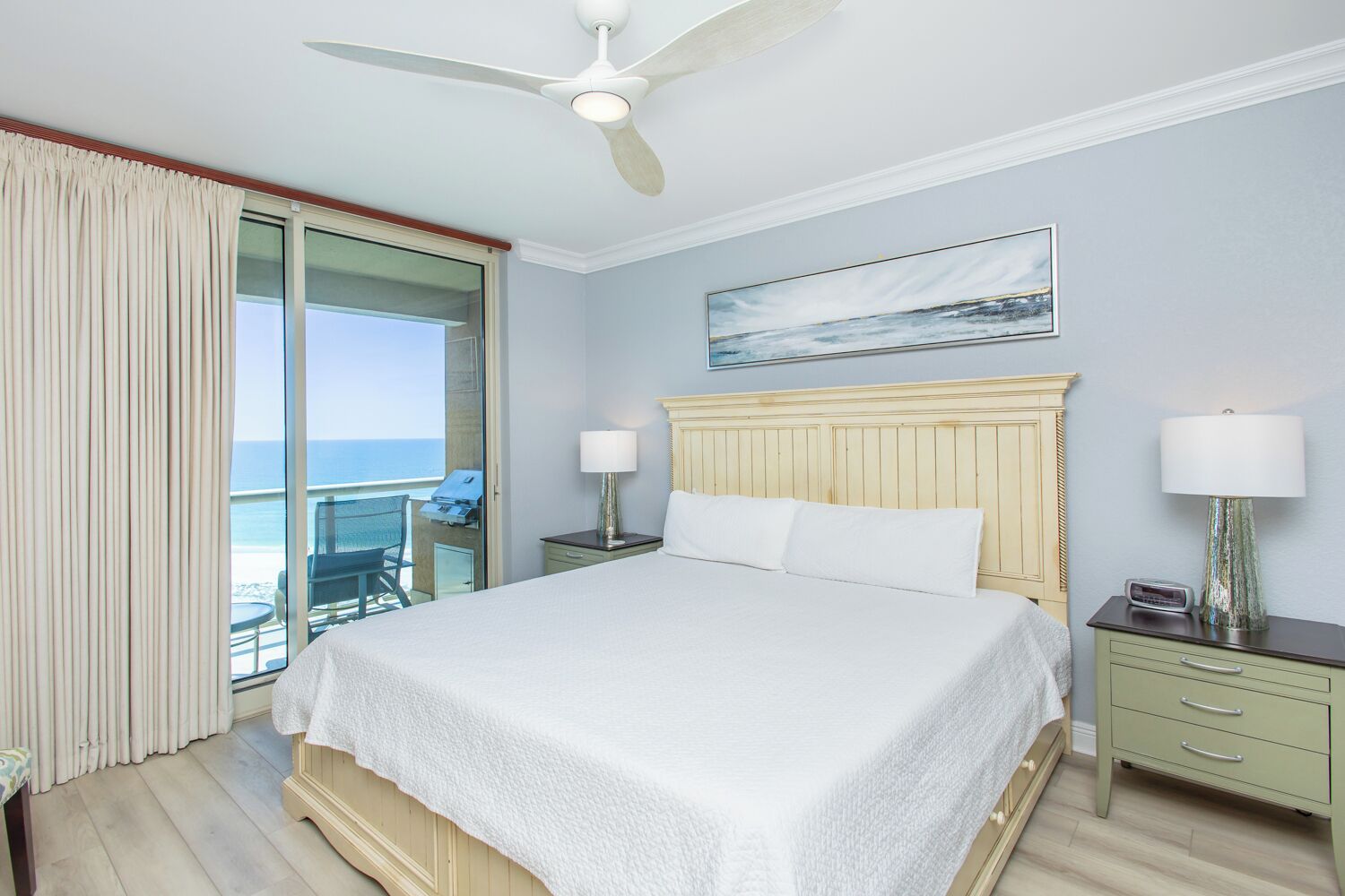 Une chambre d’hôtel avec un lit, deux tables de chevet, un ventilateur de plafond, un balcon avec une chaise et un tableau au mur.