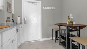 Une cuisine avec une porte blanche, un comptoir en marbre, une table en bois sur laquelle se trouvent une bouteille et une fleur, et un luminaire fixé au mur.