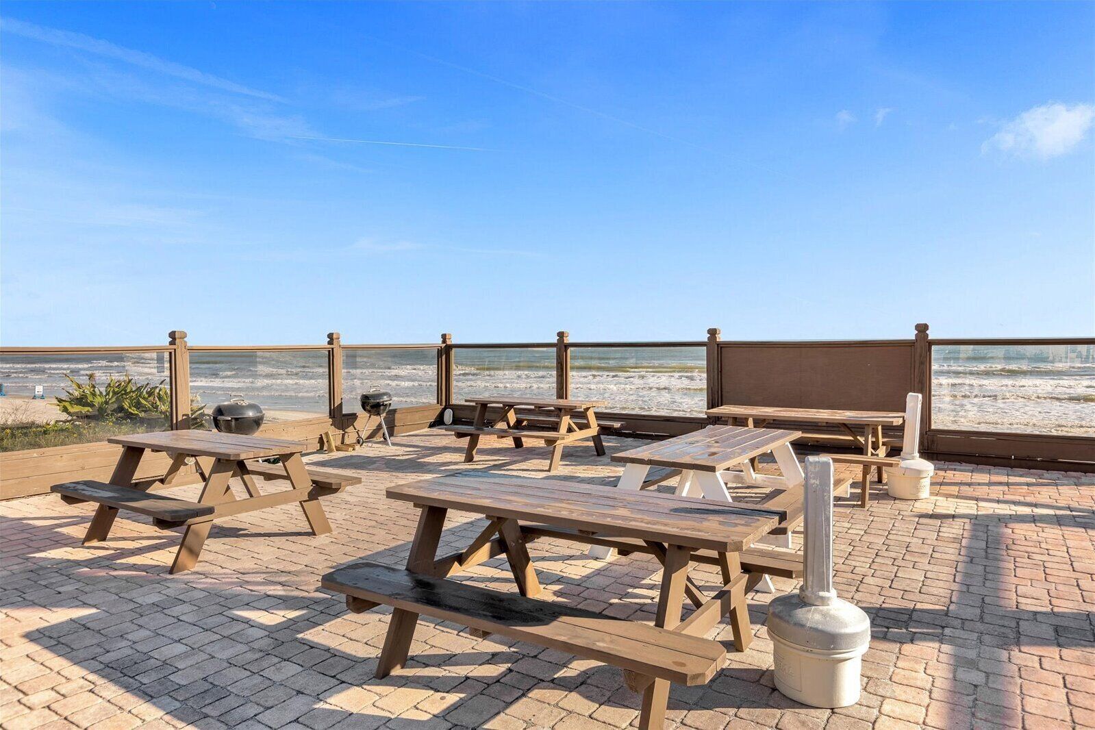 A beachside patio with picnic tables and a barbecue grill, overlooking the ocean.
