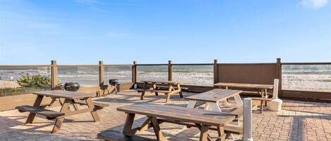 A beachside patio with picnic tables and a barbecue grill, overlooking the ocean.
