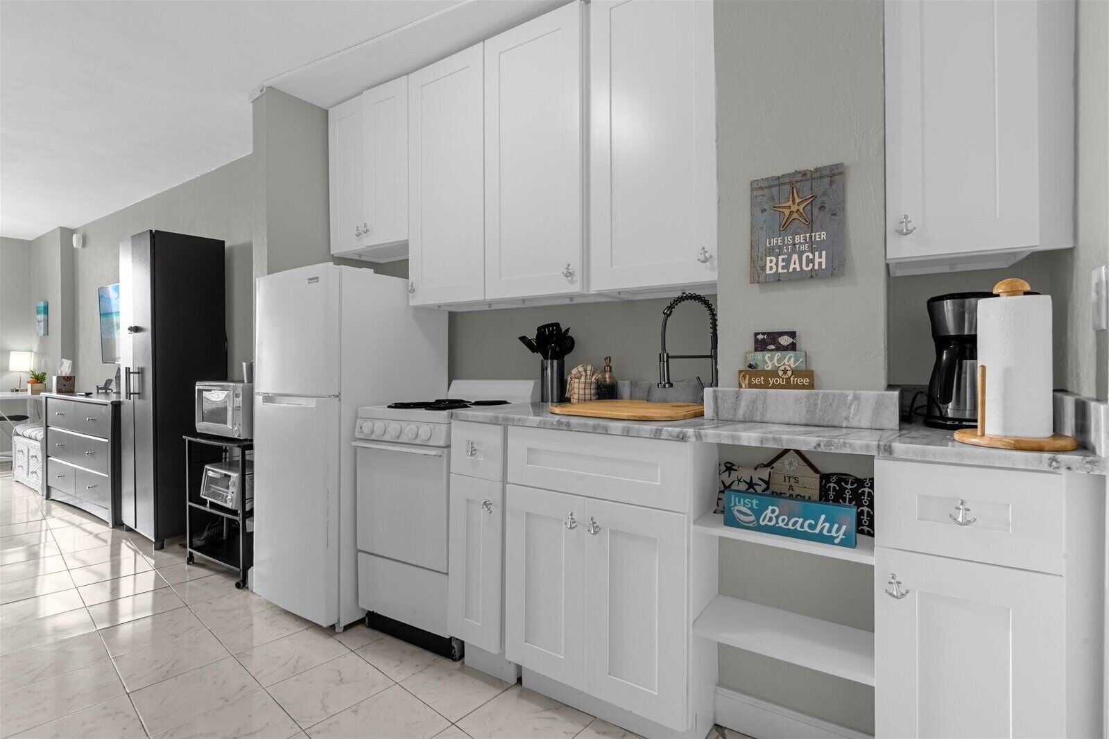 A modern kitchen with white cabinetry, a stove, and a coffee maker. There are decorative items on the counter and a sign on the wall that says "Life is better at the beach."