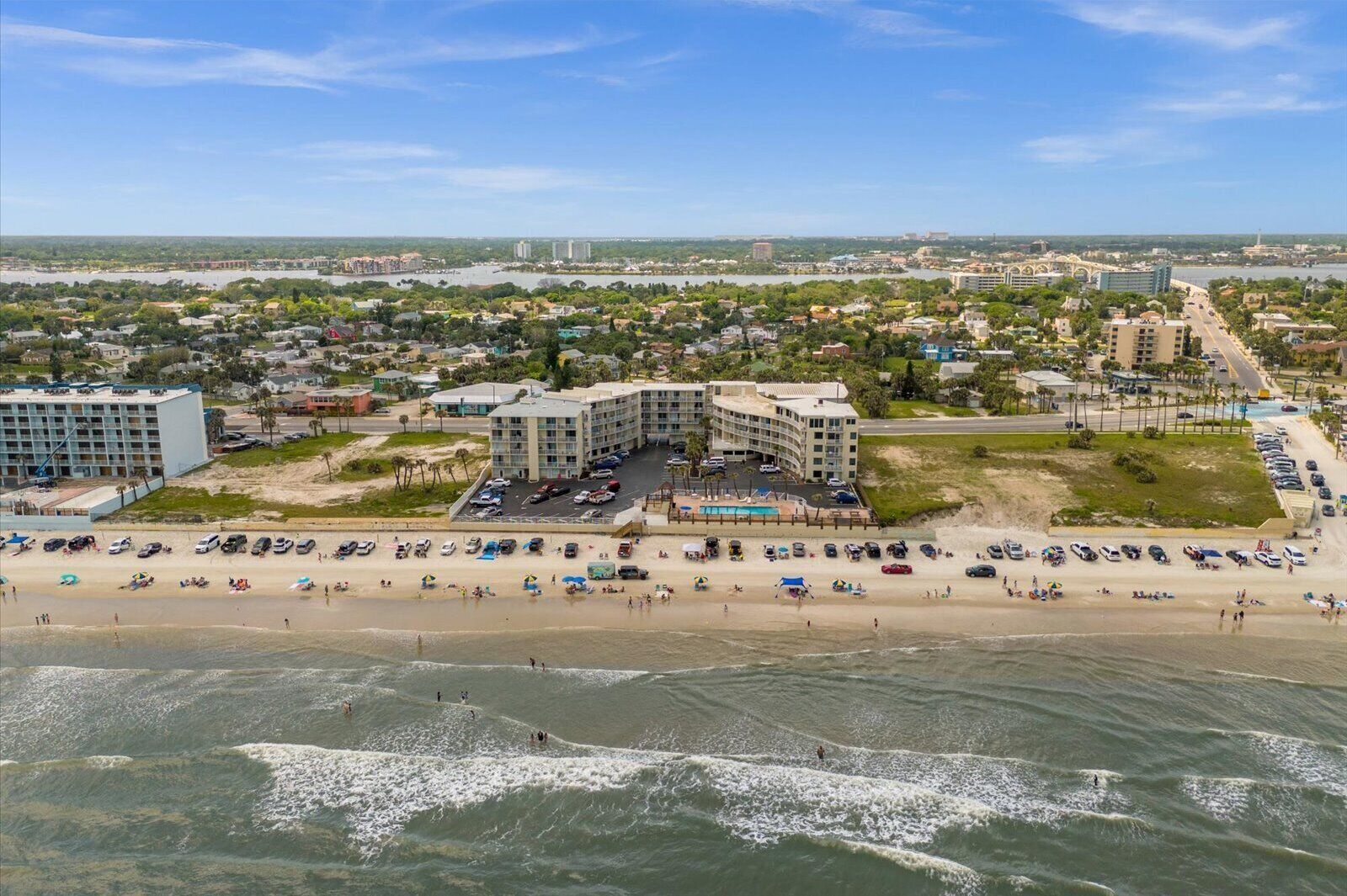 Vue aérienne d’un hôtel situé sur la côte, composé de plusieurs bâtiments, avec une plage et un parking.
