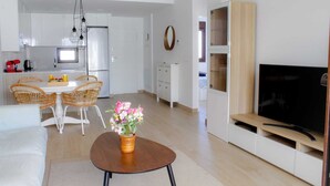Un salon moderne avec un canapé, une table basse ornée d’un vase de fleurs, un coin repas avec des chaises en osier, une cuisine avec des meubles blancs et un téléviseur posé sur un meuble en bois.