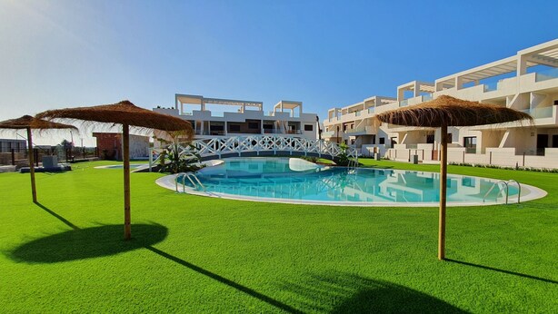 Un hôtel moderne doté d’une piscine centrale, entourée de gazon synthétique et de parasols en chaume.